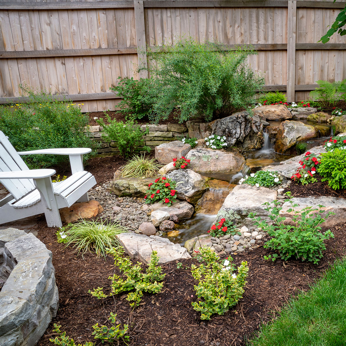 A small pondless waterfall and stream in a backyard garden beside an Adirondack chair