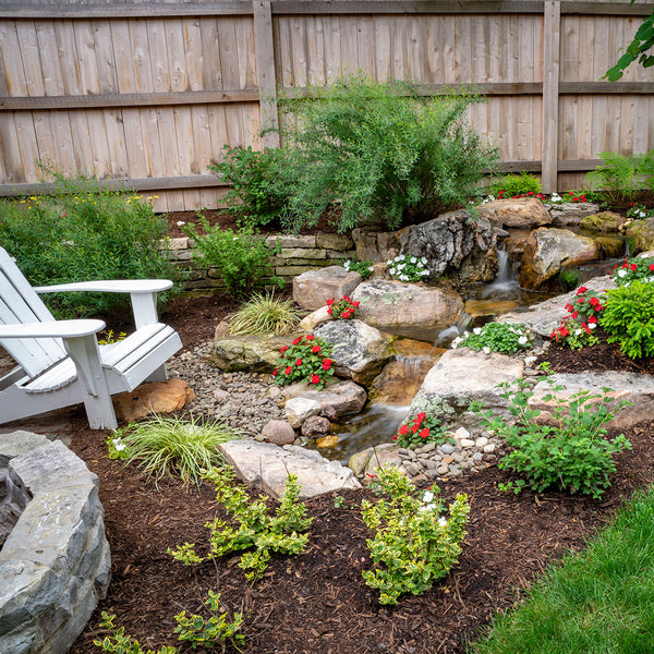 A small pondless waterfall and stream in a backyard garden beside an Adirondack chair
